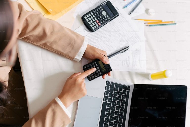 Pessoa usando calculadora sobre documentos, com um notebook ao lado
